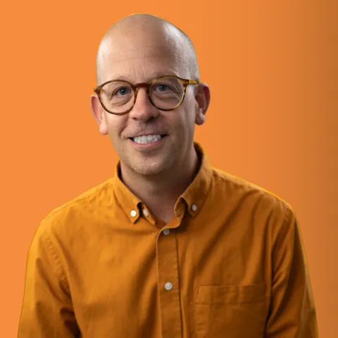 Kevan smiling at the camera, in glasses and a mustard shirt, against orange